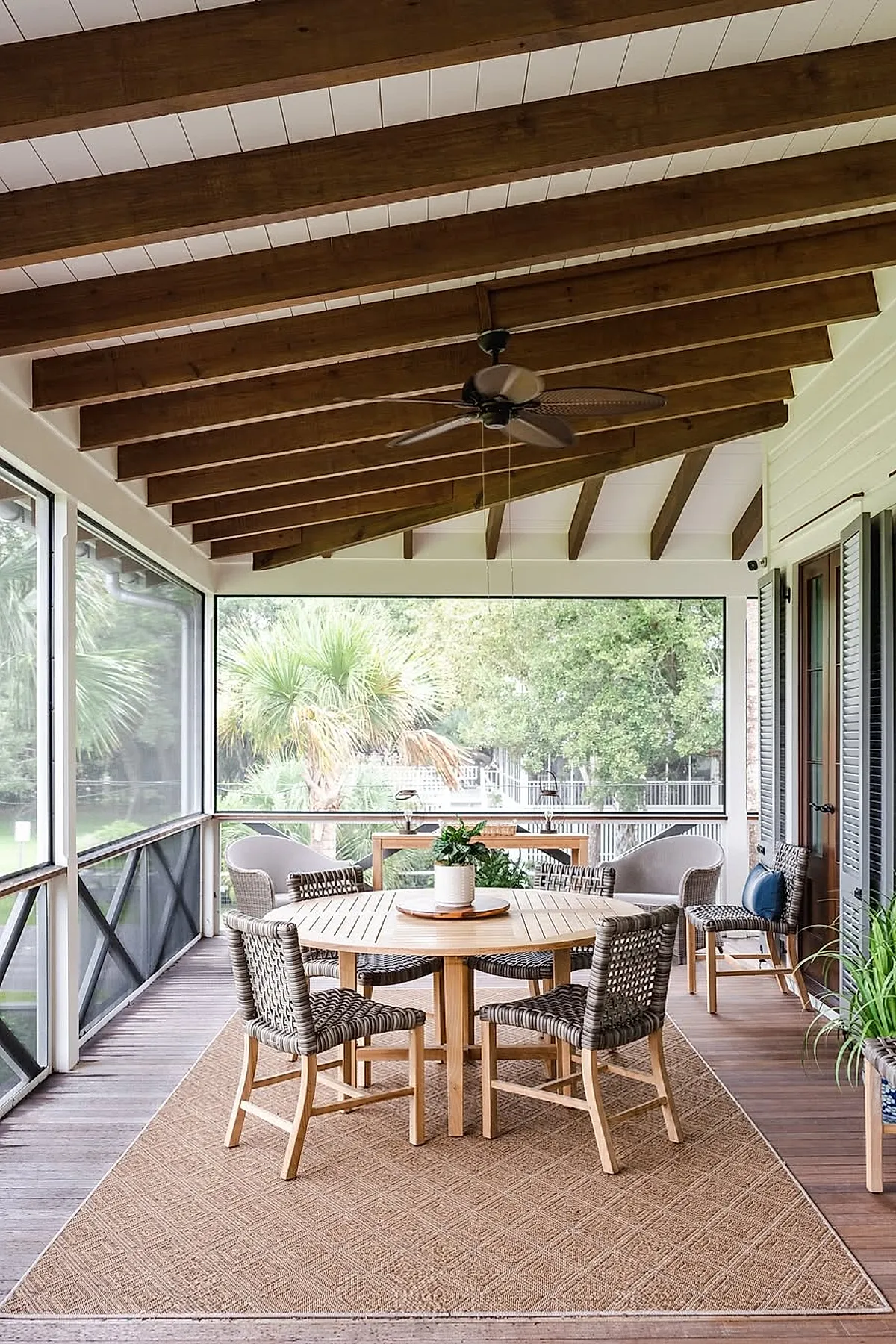 Screened in porch featuring exposed wooden beam ceiling, round wooden dining table with woven chairs, textured area rug, large screened windows showing green trees outside