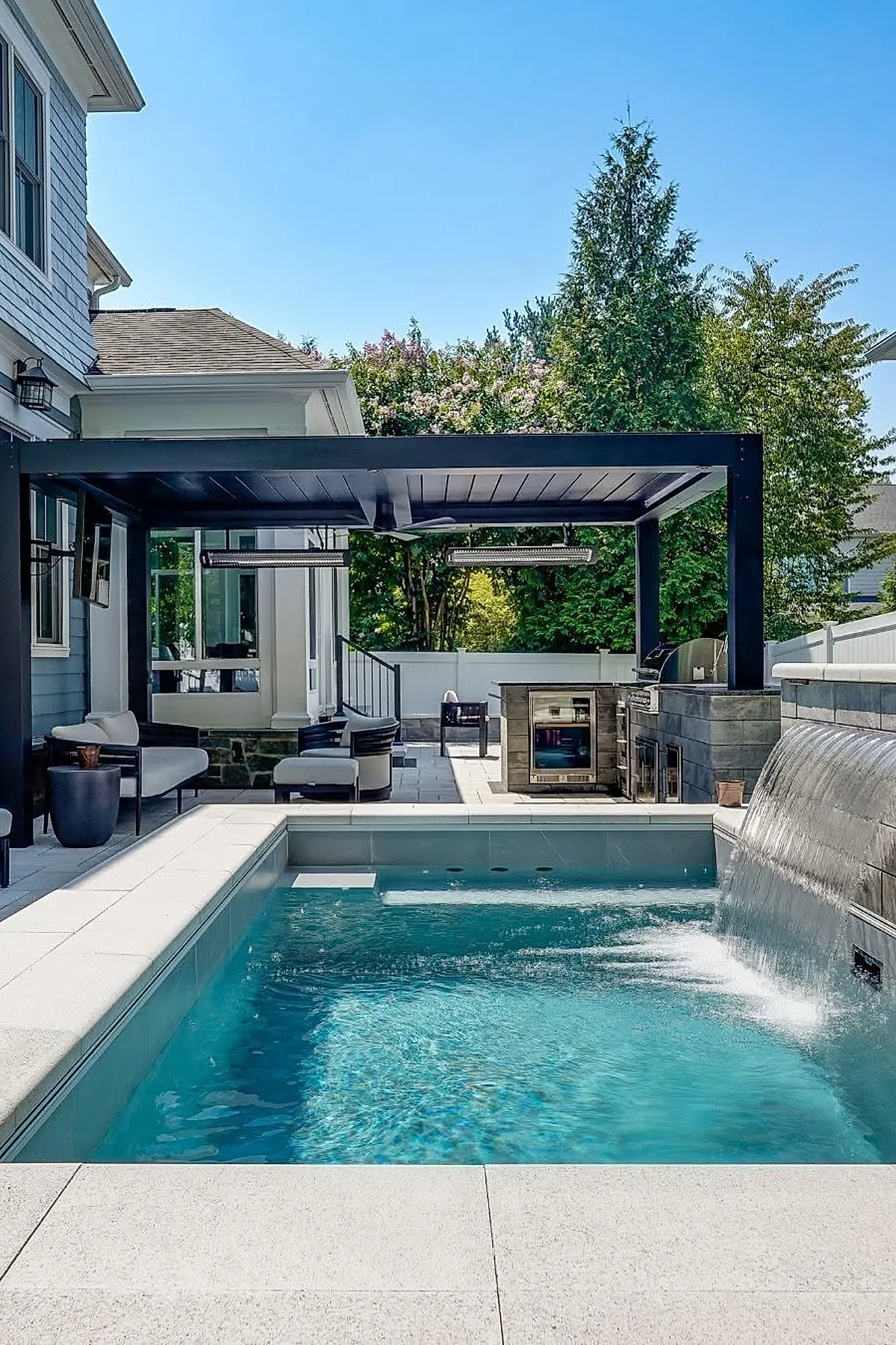 Small rectangular inground pool with clear blue water and a waterfall feature on one side, surrounded by light gray stone tiles, adjacent to a modern outdoor kitchen with grill and seating area under a black pergola, set in a backyard with green trees and blue sky.