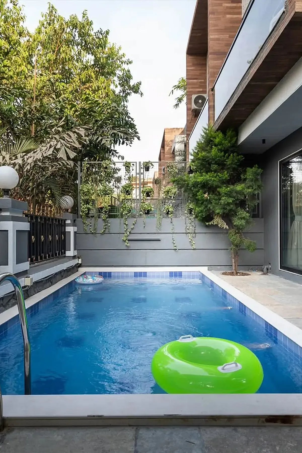A small inground pool with blue tiles surrounded by a concrete deck and modern building facade, accented by green plants and a bright green inflatable ring floating on the water.