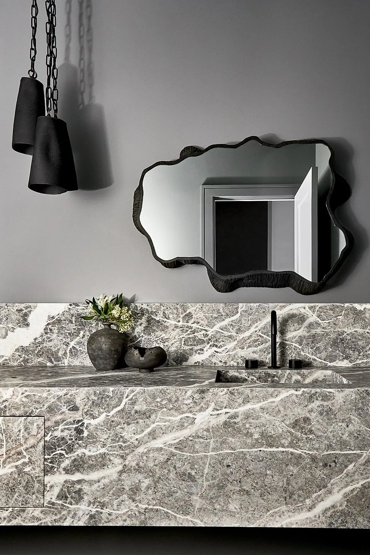 Bathroom featuring an organically shaped large mirror with a textured dark frame above a gray and white marble vanity countertop and backsplash, complemented by black pendant lights and rustic decor pieces.