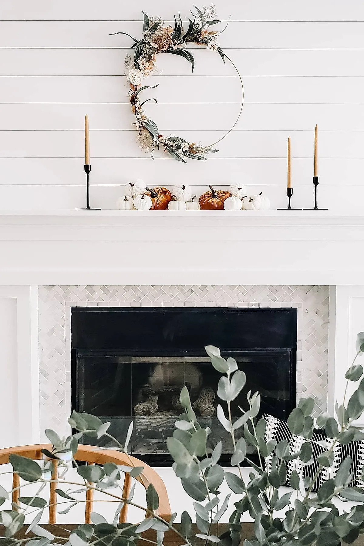 white shiplap wall above a white mantel decorated with clustered small white and orange pumpkins centered between three tall black candle holders each with beige tapered candles, above hangs an open circular wreath decorated asymmetrically on one side with dried flowers and eucalyptus leaves