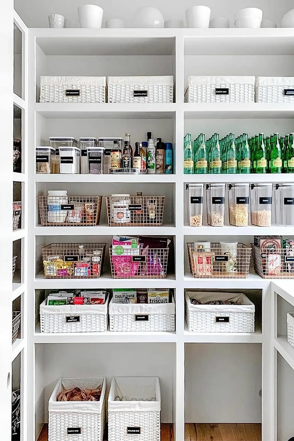White built-in pantry shelves filled with white woven baskets labeled for various items, wire baskets holding snacks and groceries, clear plastic containers with dry goods, and a row of green glass bottles neatly arranged on a shelf.
