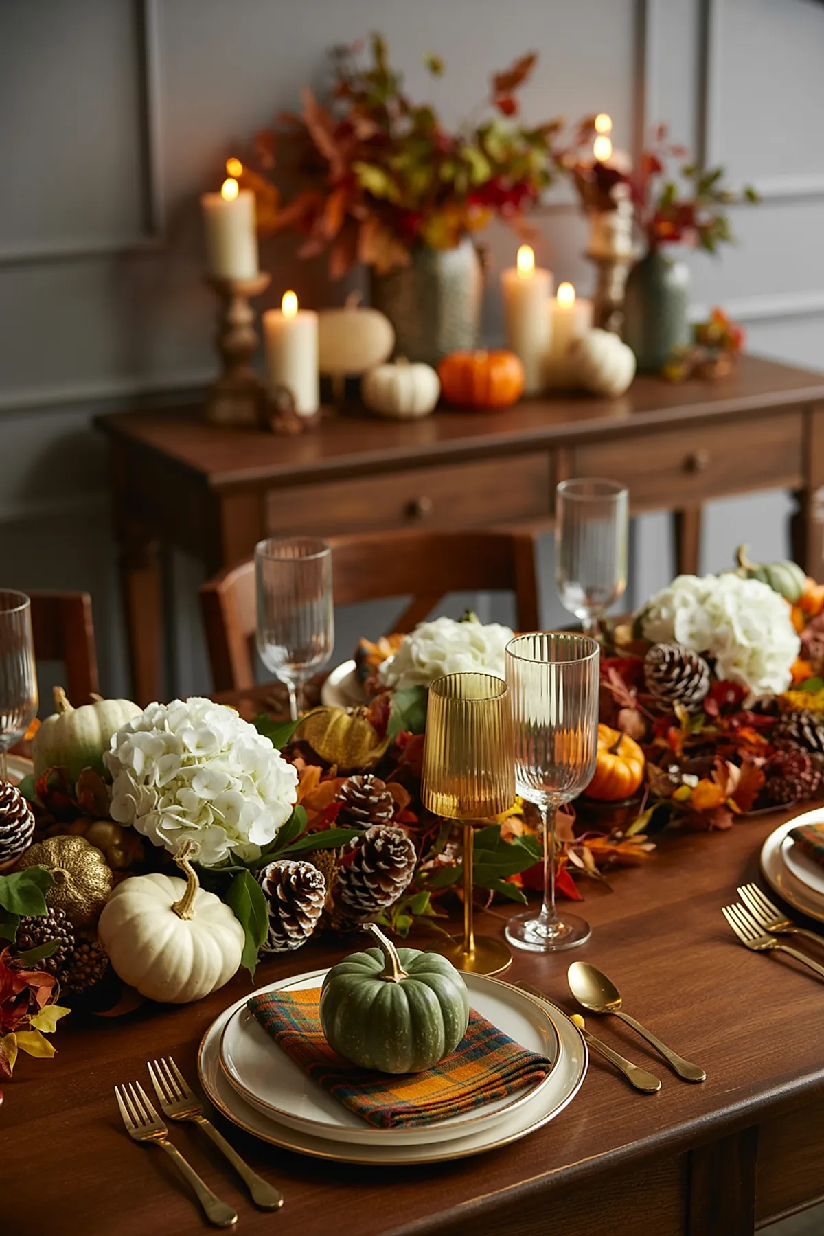Thanksgiving table with wooden surface decorated with white hydrangea flowers, brown pinecones, small white, green, and orange pumpkins, plaid napkins under pumpkins at each plate, gold flatware, amber and clear ribbed glasses, lit pillar candles on wooden console in background