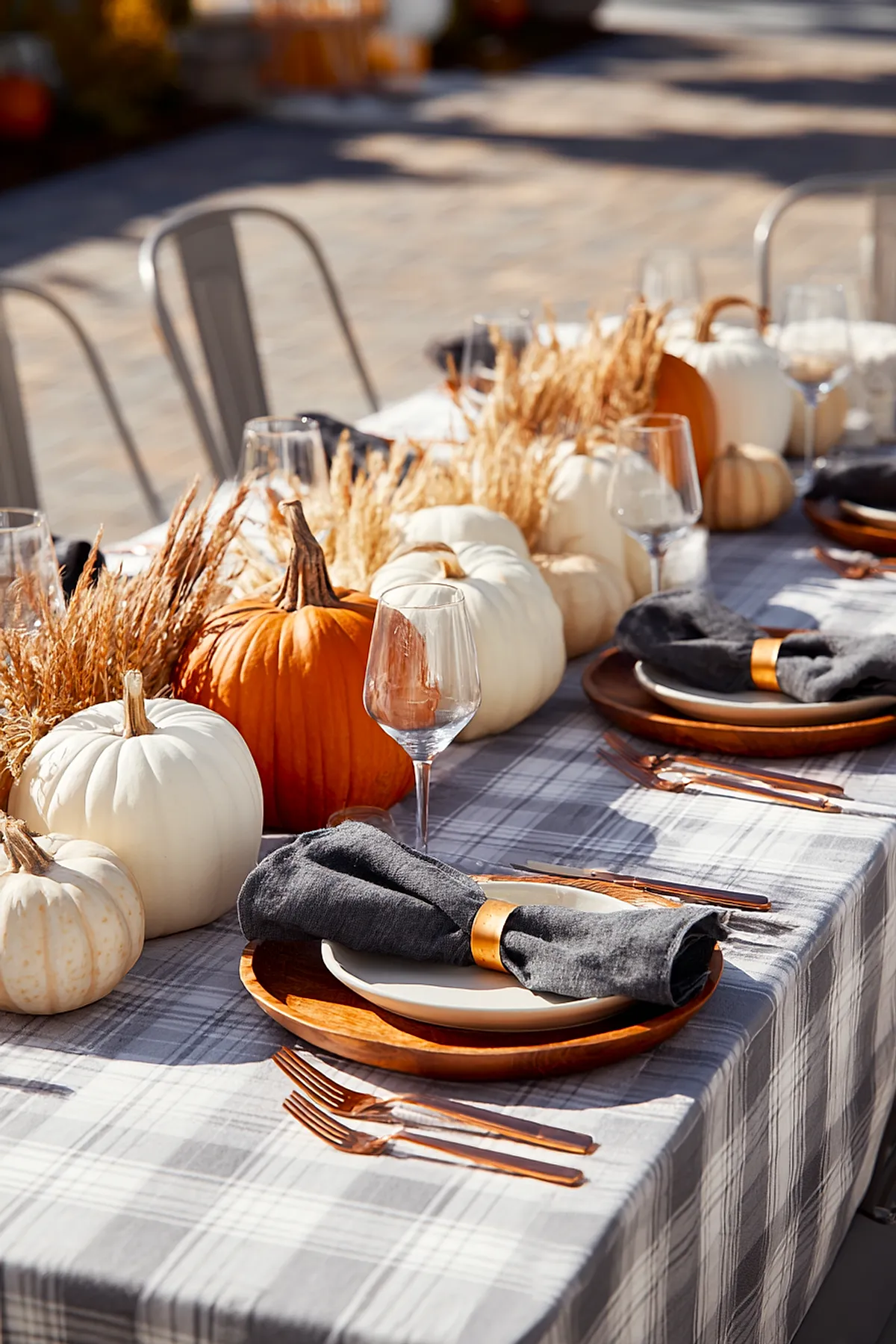 Outdoor dining table set with gray plaid tablecloth featuring a centerpiece made of white and orange pumpkins interspersed with dried wheat stalks. Each place has wooden chargers, cream plates, dark gray napkins in copper rings, clear wine glasses, and copper flatware.
