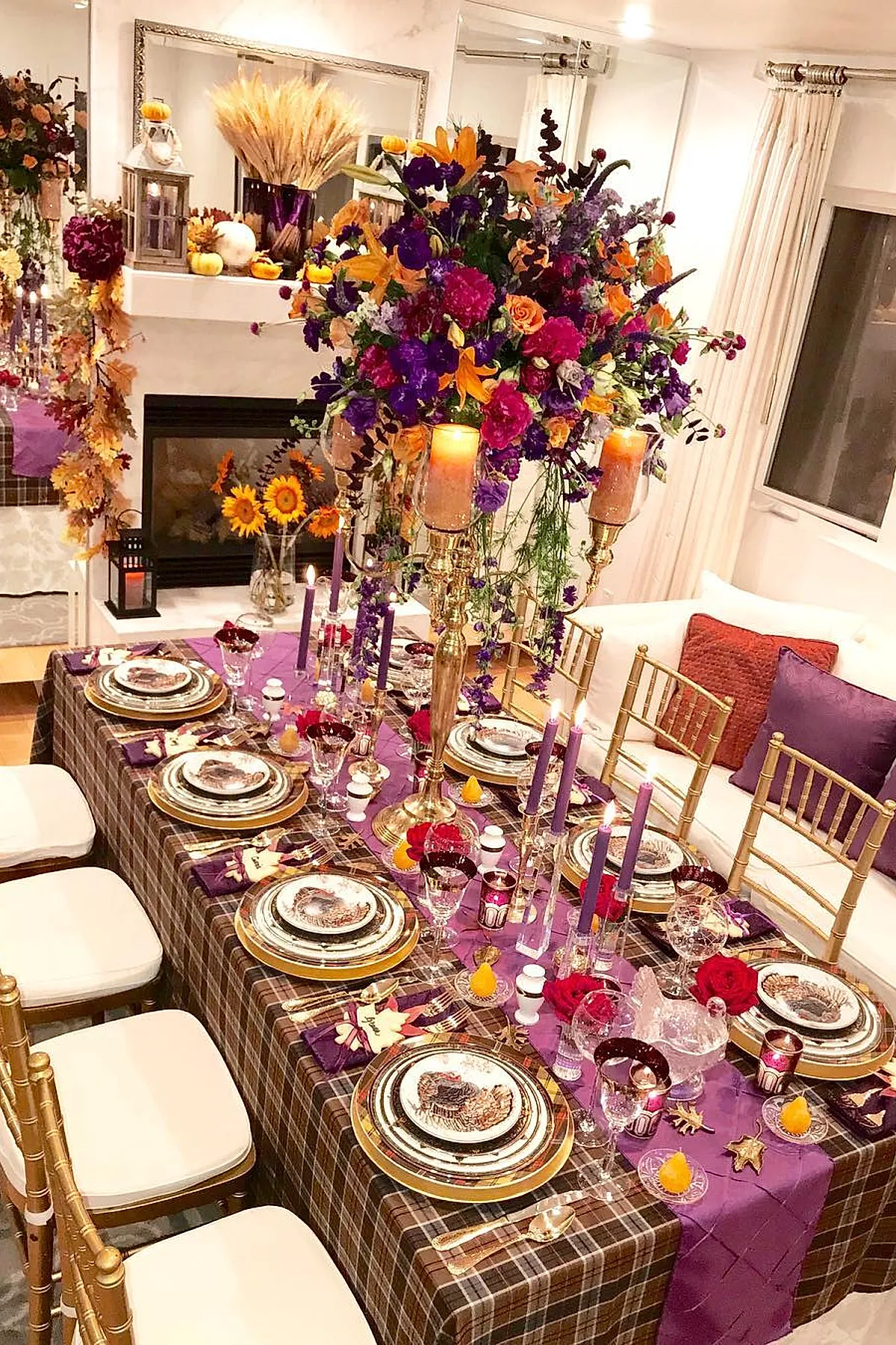 Thanksgiving dinner table with brown plaid tablecloth, purple fabric runner, tall gold candelabra holding lit candles surrounded by vibrant purple, orange, pink flowers; gold-rimmed plates with turkey design; gold flatware; purple napkins; clear crystal glasses; gold chairs with white cushions