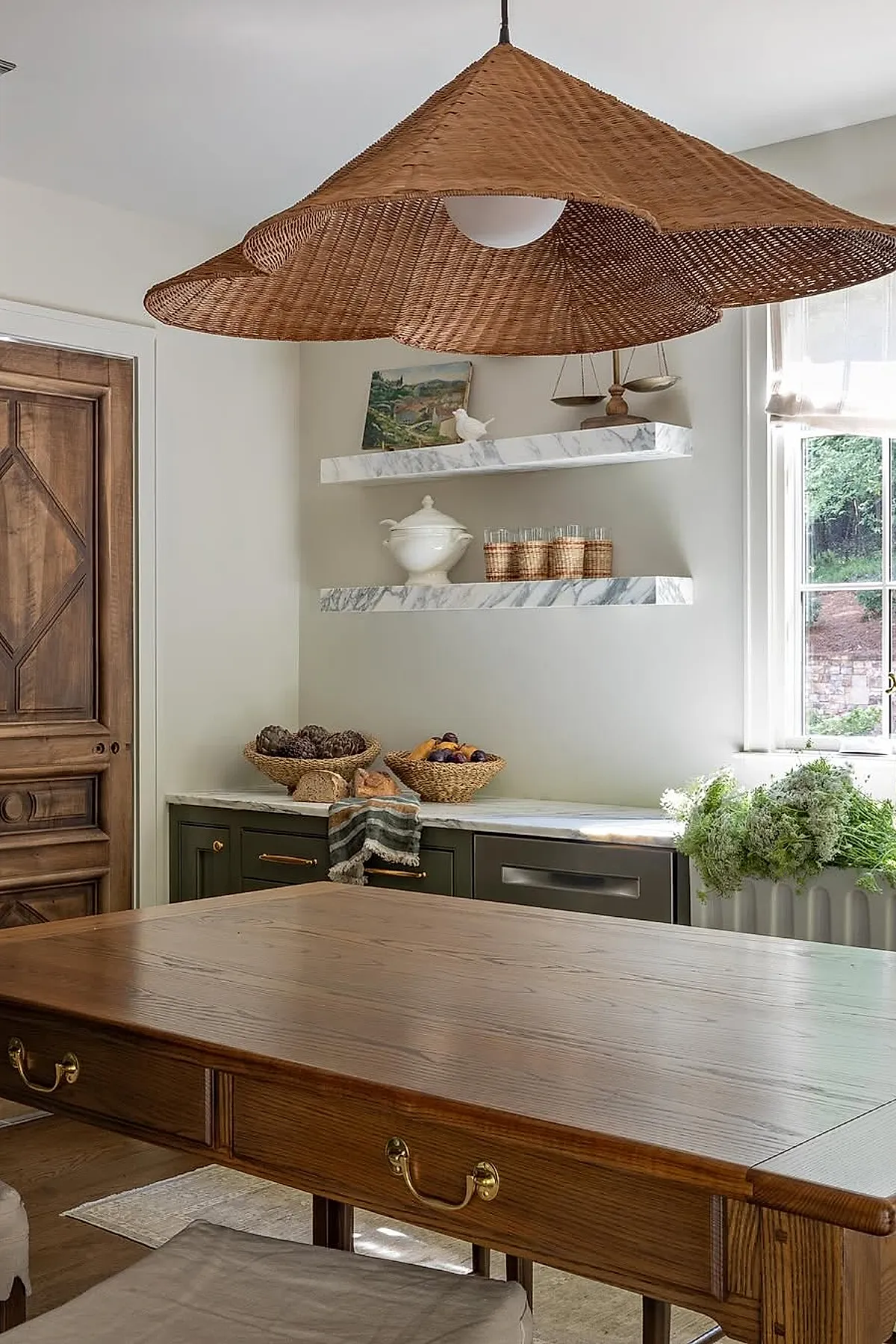 Rustic kitchen corner featuring dark green cabinetry with brass handles, white marble countertop and floating marble shelves holding wicker glasses, ceramic dish, small painting, and decorative scales, complemented by woven baskets on the counter and a large rattan pendant light overhead.
