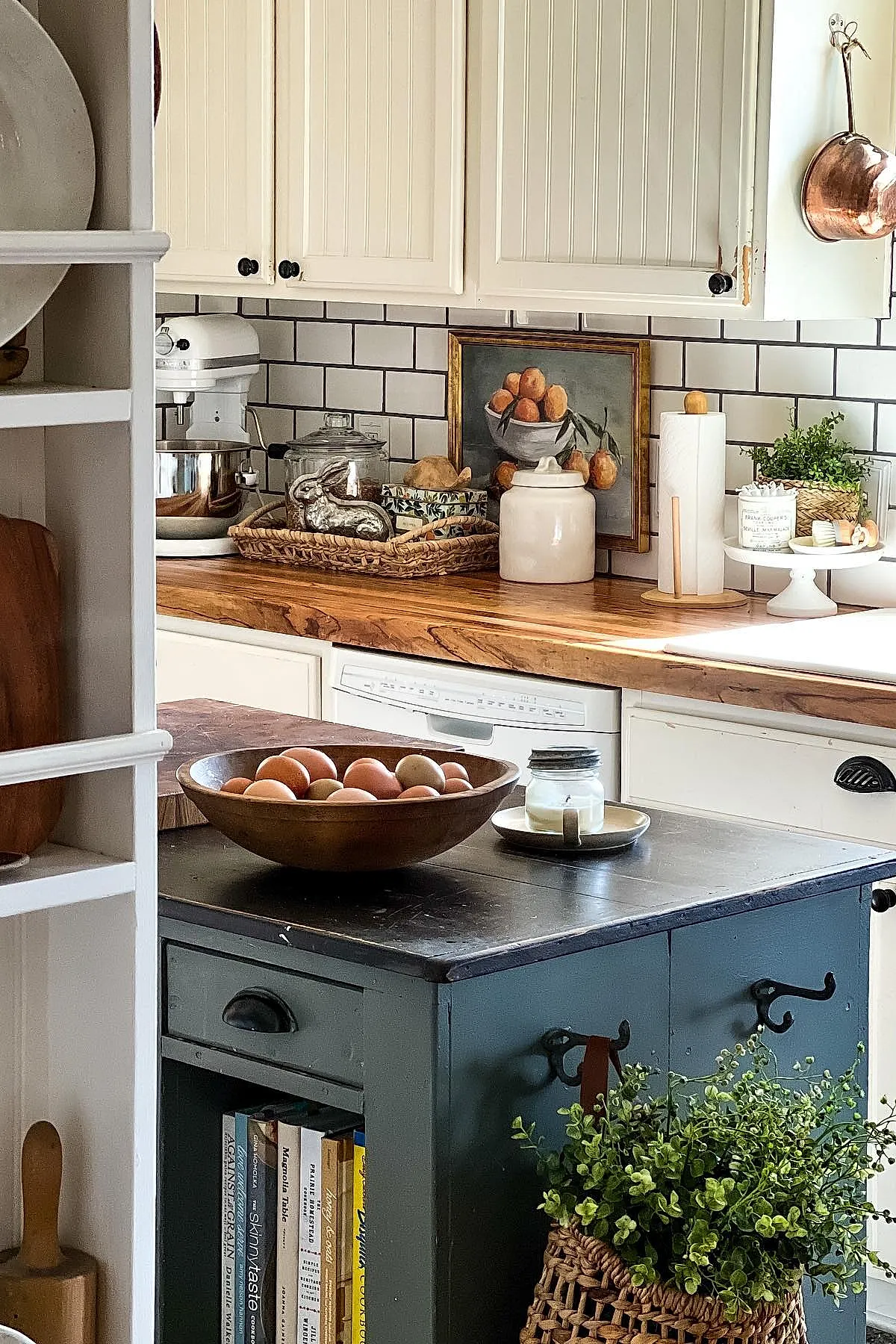Vintage cottage kitchen featuring white paneled cabinets with black knobs, wooden countertop with natural grain, white subway tile backsplash with dark grout, a white stand mixer, wicker tray holding decorative items including a silver bunny figurine, paper towel holder with wooden accents, framed fruit painting leaning against the wall, potted plant in woven basket on a pedestal cake stand, and a bowl of brown eggs on a dark blue kitchen island with books stored underneath.