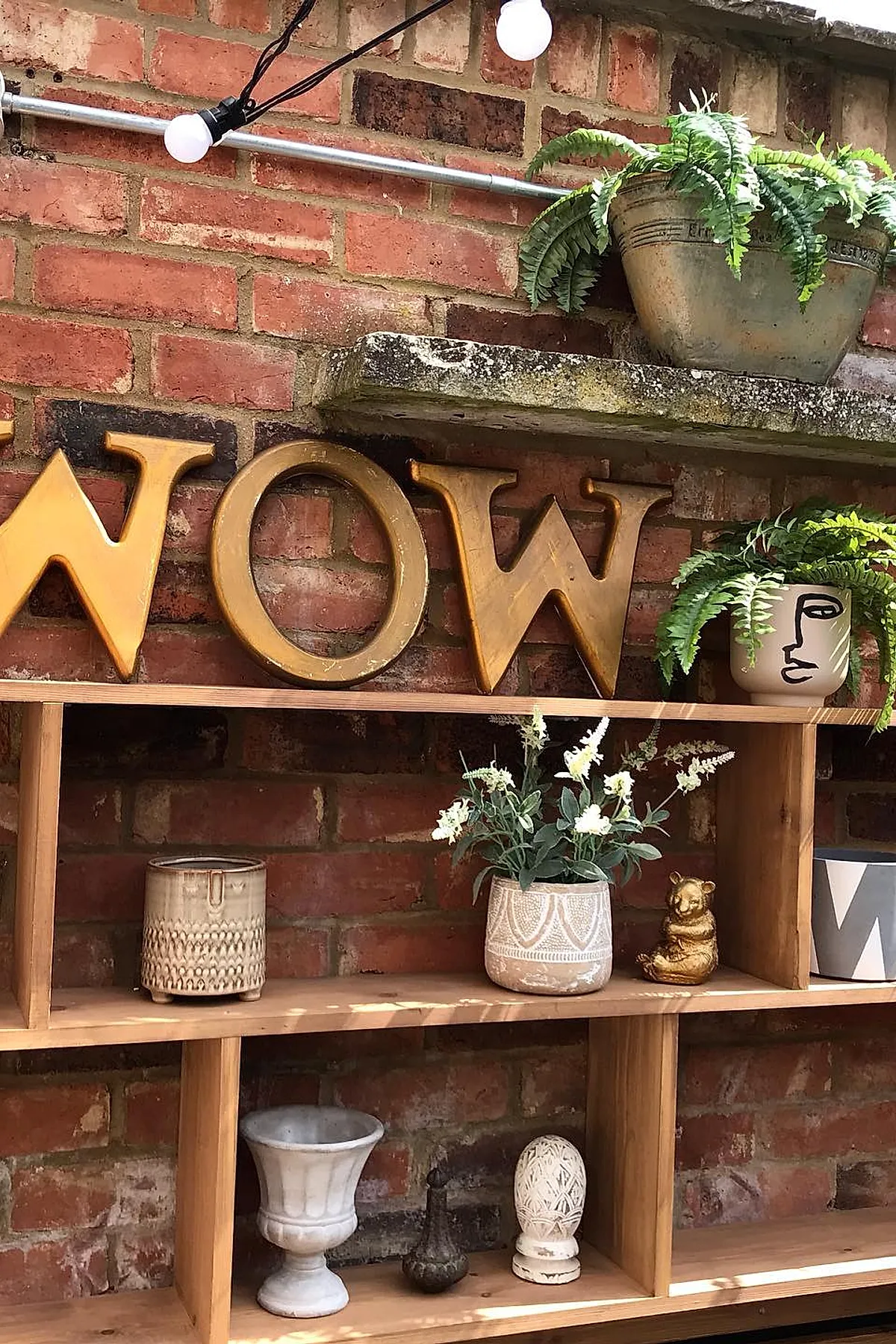 Wooden outdoor plant shelf holding decorative ceramic pots, ferns in a pot with face design, small golden bear figurine, and white flowering plants against a red brick wall