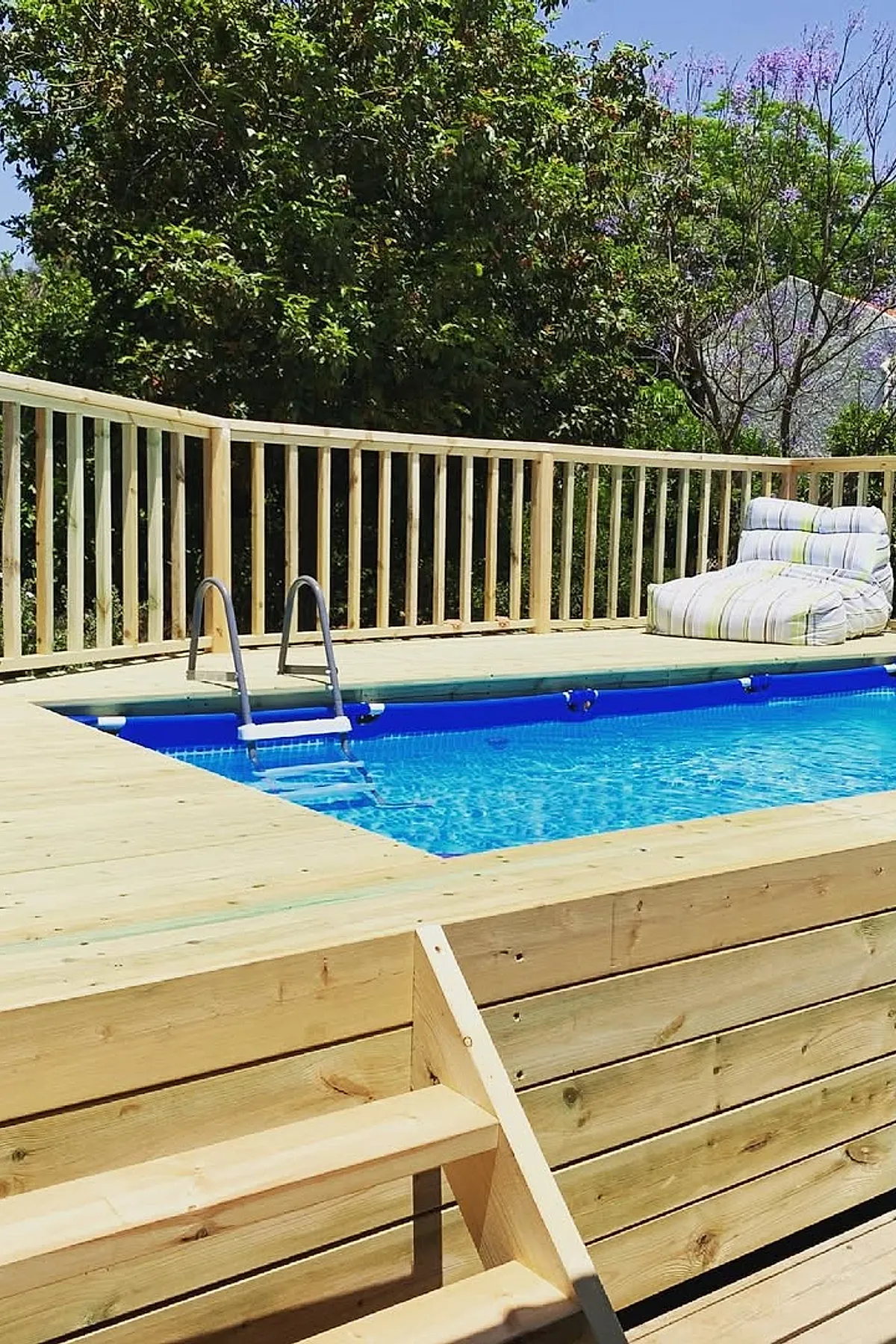 Above-ground pool with bright blue water surrounded by a natural wood pool deck featuring wooden railing and a cushioned lounge chair with white and green stripes, set against a backdrop of leafy trees under clear blue sky.