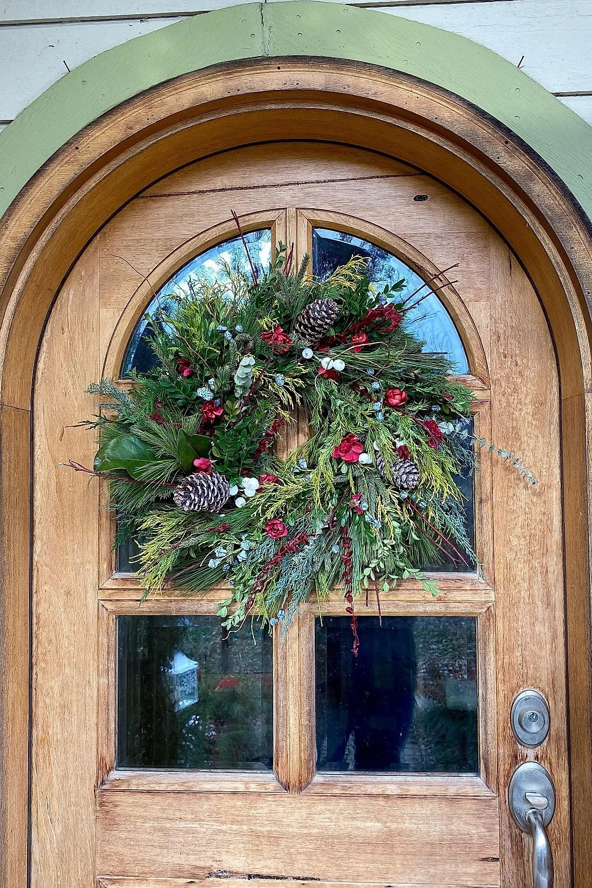 Christmas wreath featuring mixed evergreens, red berries, pinecones, and blue-gray accents hung on a wooden door with arched glass panes.