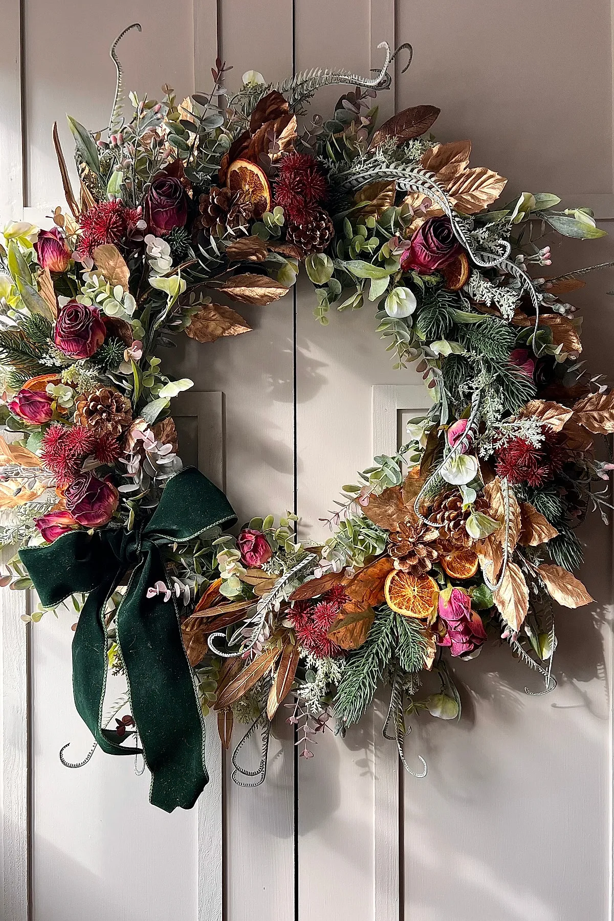 Christmas wreath featuring dried orange slices, pinecones, burgundy roses, assorted greenery, brown leaves, and a dark green velvet bow hanging on a beige door.