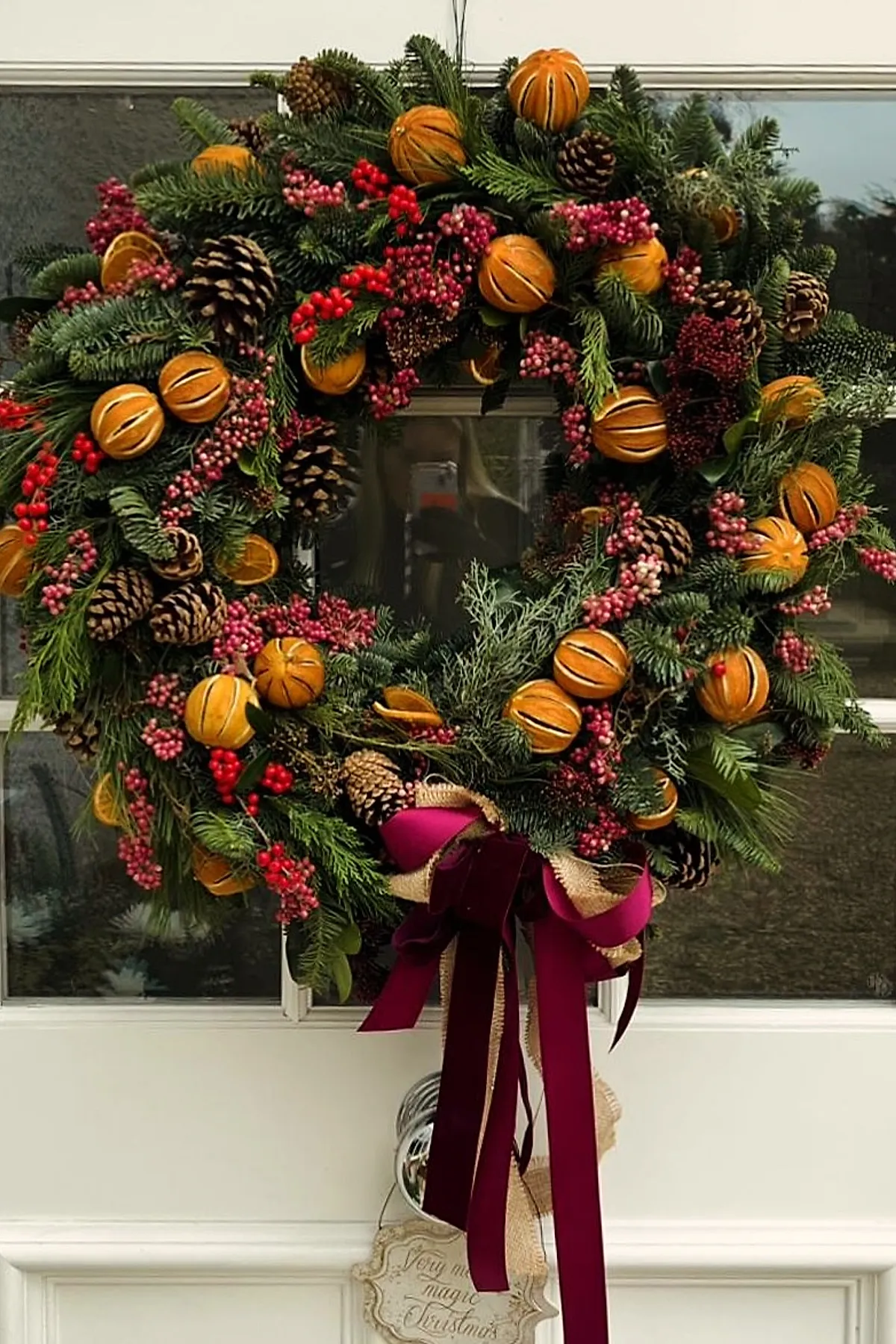 Christmas wreath made of lush evergreen branches decorated with dried orange pomanders, clusters of red and pink berries, pinecones, and tied with burgundy velvet and burlap ribbons hanging on a white door.