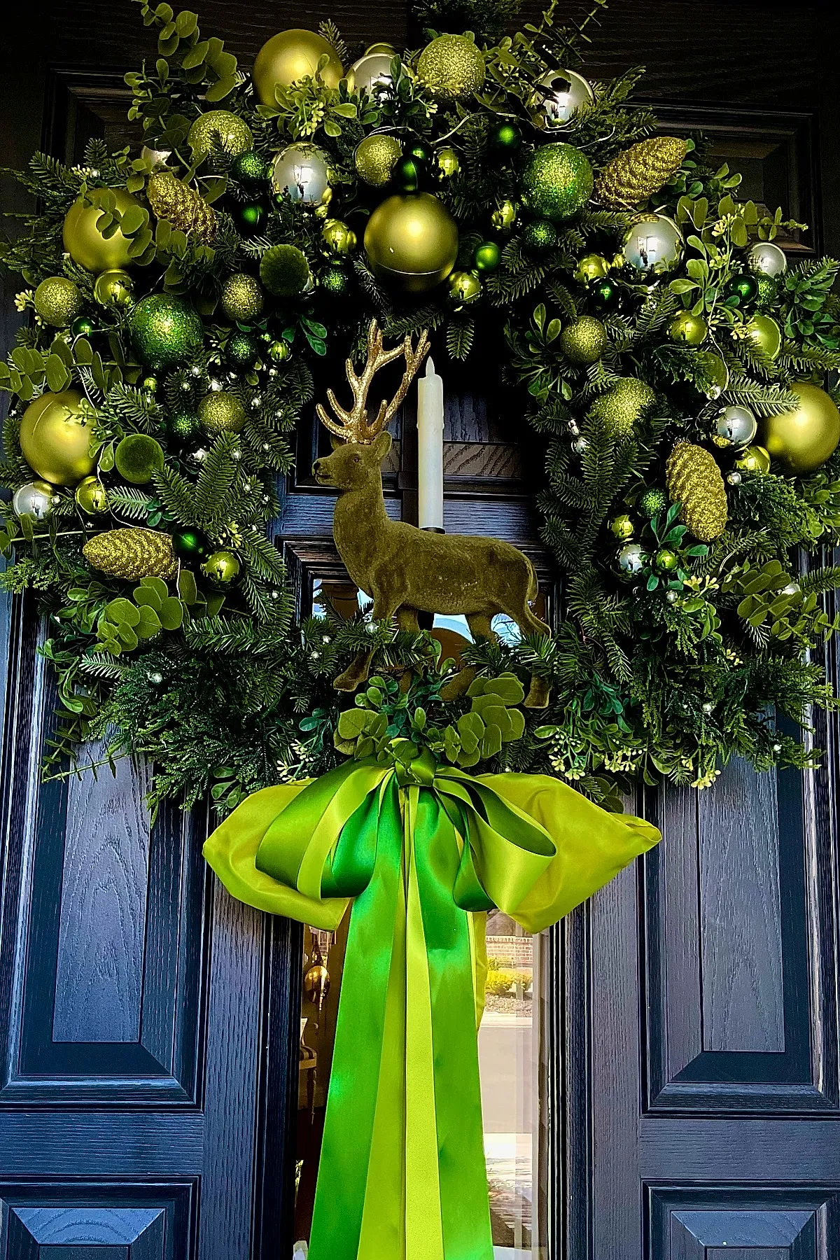 Christmas wreath decorated with green foliage, assorted green and gold ornaments, glittery pinecones, a velvet reindeer figure in the center, and large two-tone green satin bow hanging on a dark door.