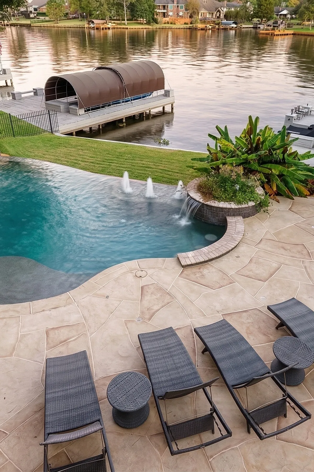Curved pool deck made of beige stone tiles featuring water fountains and a waterfall flowing into the blue swimming pool, surrounded by gray wicker lounge chairs and green plants with a lake and dock visible in the background.